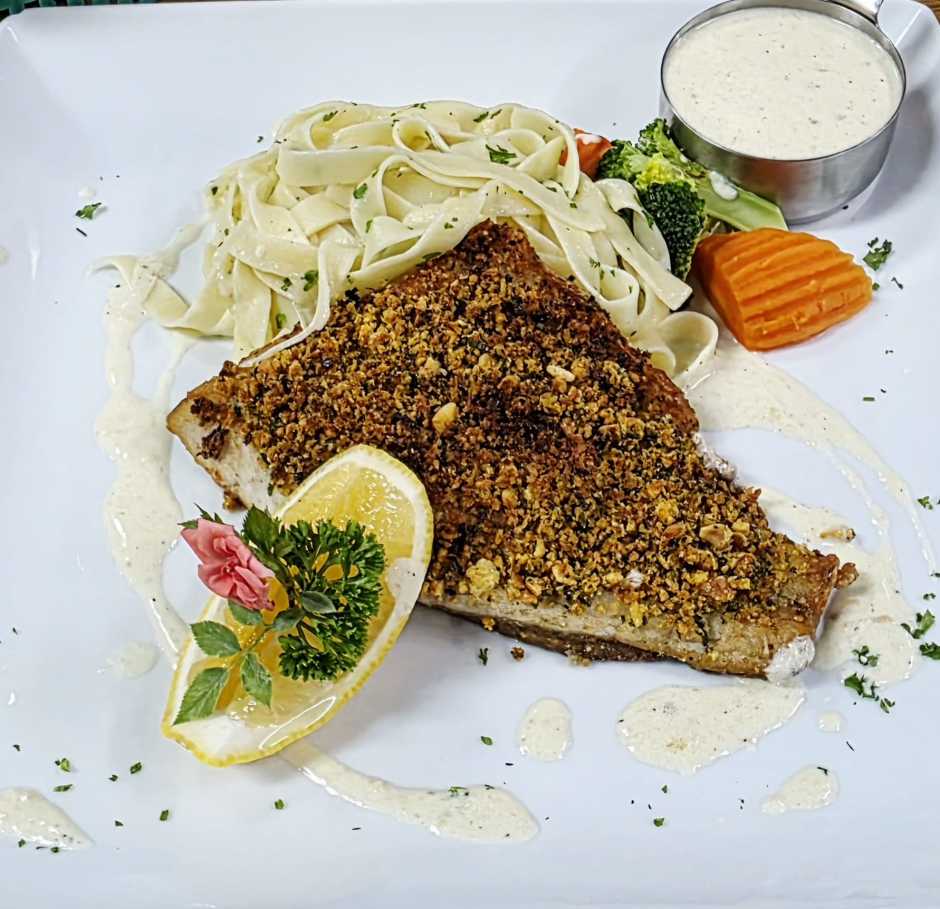 Das Wolfsbarschfilet in Kräuterkruste wird mit Parmesan-Rahmsauce, Bandnudeln und Buttergemüse serviert.